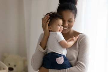 Wall Mural - Front view affectionate african american mum holding little funny mixed race toddler daughter or son, enjoying spending daycare time together in baby room, showing love and devotion to small kid.