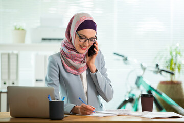 Wall Mural - Beautiful young working woman in hijab and suit sitting in office, using smart phone. Portrait of confident muslim businesswoman. Modern light office with big window. 