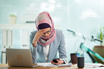 Poster - Beautiful young working woman in hijab, suit and eyeglasses sitting in office, having headache. Modern light office with big window. 