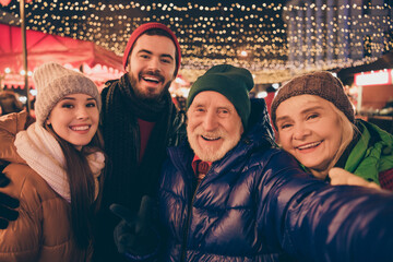 Closeup portrait photo of full family four relatives people x-mas meeting take selfie show v-sign hug outerwear hat scarf coat two generation evening street illumination outdoors outside