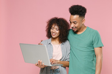 Smiling young african american couple two friends guy girl in gray green casual clothes posing hugging working on laptop pc computer isolated on pastel pink color wall background studio portrait.