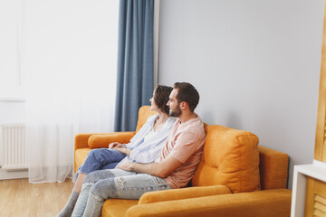 Wall Mural - Side view of smiling attractive beautiful young couple two friends man woman 20s wearing casual clothes sitting on couch hugging looking aside resting relaxing spending time in living room at home.