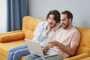 Canvas Print - Shocked amazed excited young couple two friends man woman in casual clothes sitting on couch hugging pointing index finger on laptop pc computer resting relaxing spending time in living room at home.