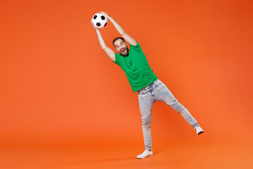 Poster - Full length portrait excited young man football fan in green t-shirt cheer up support favorite team catching soccer ball isolated on orange background studio. People sport leisure lifestyle concept.