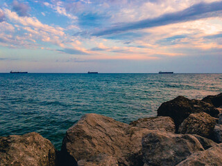 Wall Mural - The surface of the sea against a blue cloudy sky with three ships on the horizon and large stones in the foreground.