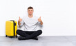 © luismolinero - Young handsome man sitting on the floor with a suitcase giving a thumbs up gesture