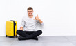 © luismolinero - Young handsome man sitting on the floor with a suitcase making phone gesture