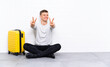 © luismolinero - Young handsome man sitting on the floor with a suitcase smiling and showing victory sign