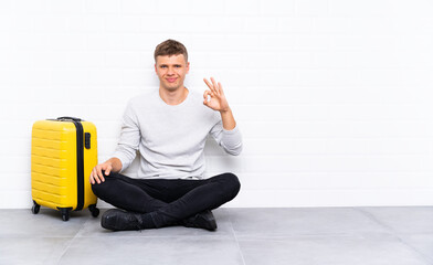 Wall Mural - Young handsome man sitting on the floor with a suitcase showing an ok sign with fingers