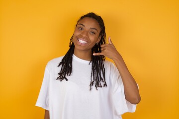 Wall Mural - Young dark skinned woman with braids hair wearing white t-shirt over yellow background smiling doing phone gesture with hand and fingers like talking on the telephone. Communicating concepts.