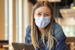 Young woman sitting at coffee shop and working on digital tablet with face mask
