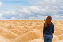A Young Woman With Red Hair Stands With Her Back To The Camera In A Desert With Sand Dunes Of Bizarre Shape