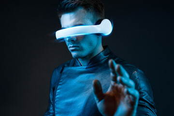 Young man in futuristic costume on dark background. Guy using VR helmet. Augmented reality, virtual reality, future technology, game concept. Blue neon light. 