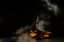 Three Jack O Lanterns Glow In The Dark Amidst The Fog. Halloween Pumpkin In A Witch Hat.