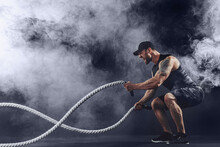 Bearded Athletic Looking Bodybulder Work Out With Battle Rope On Dark Studio Background With Smoke. Strength And Motivation.