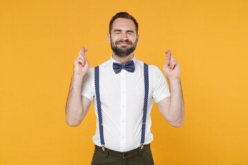 Young bearded man 20s wearing white shirt bow-tie suspender posing waiting for special moment keeping fingers crossed eyes closed making wish isolated on yellow color wall background, studio portrait.