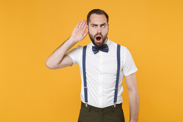 Shocked curious young bearded man 20s wearing white shirt bow-tie suspender posing try to hear you overhear listening intently looking camera isolated on yellow color wall background studio portrait.