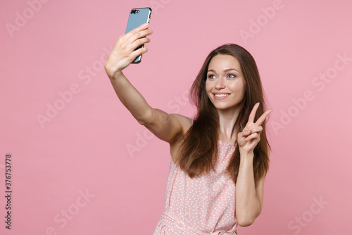 Fotografía Smiling beautiful young brunette woman 20s wearing pink summer dotted dress posing doing selfie shot on mobile phone showing victory sign isolated on pastel pink color wall background studio portrait