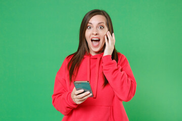 Wall Mural - Surprised young brunette woman 20s wearing bright red casual streetwear hoodie posing using mobile cell phone typing sms message put hand on cheek isolated on green color background studio portrait.