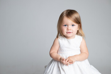 Little cute girl in a white dress. The child is 2 years old. Gently playfully smiles. Blonde hair. Blue eyes. Caucasian. Counts fingers. Portrait. Horizontal. Studio light. Grey backgroud