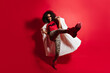 Full-length shot of adorable funny girl jumping on red background. Carefree african woman in long coat dancing in studio.