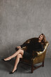© innarevyako - Happy model girl with long slim legs in a black dress sitting in a vintage leather armchair over vintage and shabby background
