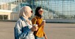 Arabian young stylish women in traditional headscarves and sunglasses walking outside at big glass building of aeroport, talking and drinking coffee. Muslim girls friends chatting and sipping drink.