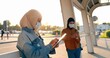 © VAKSMANV - Muslims young pretty women in traditional headscarves standing outdoors and using gadgets. Arabic females in medical masks tapping and scrolling on mobile phone and tablet device. Arabs girls in city.