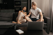© Виталий Римдейка - a beautiful laughing woman and a man are sitting on the sofa in the apartment, chatting, drinking tea and looking at something on a laptop. The relationship of a young married couple