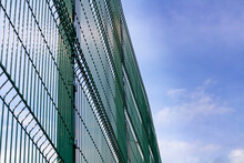 High Fence Made Of Green Steel Mesh Against A Blue Sky With Clouds. The Concept Of Protecting Property Or Territory.