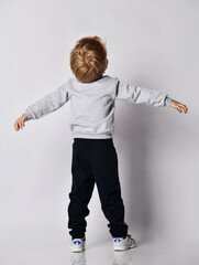 Wall Mural - Blond kid boy in black pants and sweatshirt stands back to camera holding head up and arms spread wide