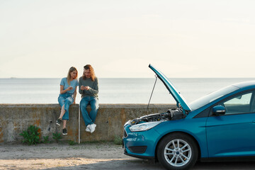 Wall Mural - Outdoor shot of couple of travelers having problems with the car, man and woman sitting near broken car with open hood and looking for a phone number of assistance, trying to solve problem