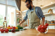 © Svitlana - Cheerful young man, Italian cook in apron putting basil leaves on the table while getting ready to prepare healthy meal with vegetables in the kitchen. Cooking at home concept
