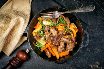 Roast pork with vegetables in a pan on a dark background, top view