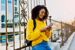 © Svetlana Sokolova - Outdoor image of young attractive black woman with stylish Afro hairs  using mobile phone and sitting on  bridge  in modern district. Yellow glasses and pullover.