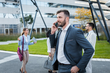 Wall Mural - Portrait of smiling businesswoman talking at  smartphone while standing in front of modern office buildings with colleagues behind him.