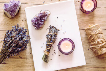 Homemade herbal lavender (lavendula) smudge stick smoldering on white plate with candles and amethyst crystal clusters for decoration. Spiritual home cleansing concept.