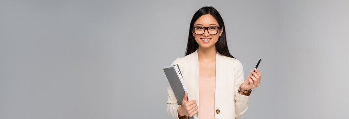 Wall Mural - panoramic shot of young asian businesswoman holding folder and pen while looking at camera isolated on grey