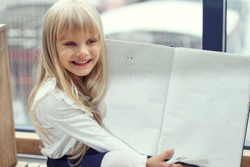 beautiful little blonde girl smiles and shows an unfolded notebook with blank sheets. layout, place for text