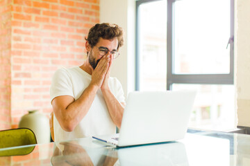 Wall Mural - young bearded man with a laptop looking happy, cheerful, lucky and surprised covering mouth with both hands