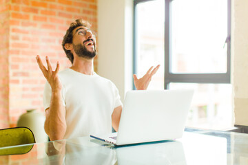 Wall Mural - young bearded man with a laptop feeling happy, amazed, lucky and surprised, celebrating victory with both hands up in the air