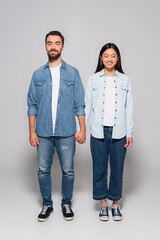 Wall Mural - full length view of interracial couple in denim clothes and gumshoes holding hands while standing with closed eyes on grey