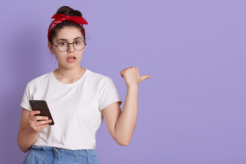 Cute young girl white t-shirt posing against lilac background, holding in hand mobile phone, pointing fingers aside on copy pace, astonished lady with cellphone.