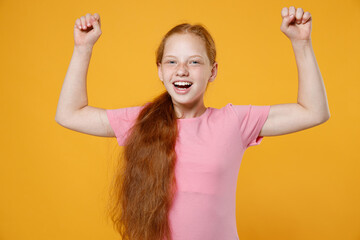 Excited little ginger redhead kid girl 12-13 years old wearing pink casual t-shirt posing clenching fists doing winner gesture isolated on bright yellow color wall background children studio portrait.