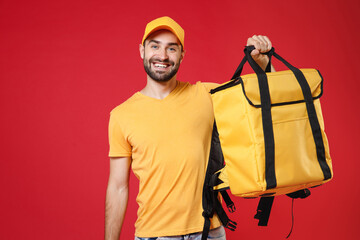 Wall Mural - Delivery employee man guy 20s in yellow cap t-shirt uniform hold thermal bag backpack with food work as courier isolated on red background studio. Service during quarantine coronavirus covid-19 virus.