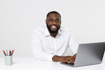 Smiling funny young african american male business man in classic white shirt posing working in office sitting at desk using laptop pc computer looking camera isolated on white color wall background.