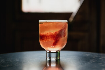 A beautiful pink cocktail with a big ice cube in a lowball glass, garnished with blood orange zest, a reflection in black marble table