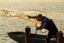 A Fisherman Wearing Overalls And Boots As Well As A Bucket Hat Is Throwing A Cast Net Into The Sea From A Wooden Pier At Sunset. He Is Backlit And Seen As Silhouette. Sun Reflects In Water.