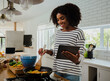 © Prins Productions - Smiling mixed race female chef reading online recipe holding digital tablet making hot curry in modern kitchen.