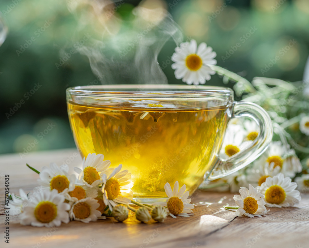 Herbal chamomile tea and chamomile flowers near teapot and tea glass on wooden table. Countryside background.